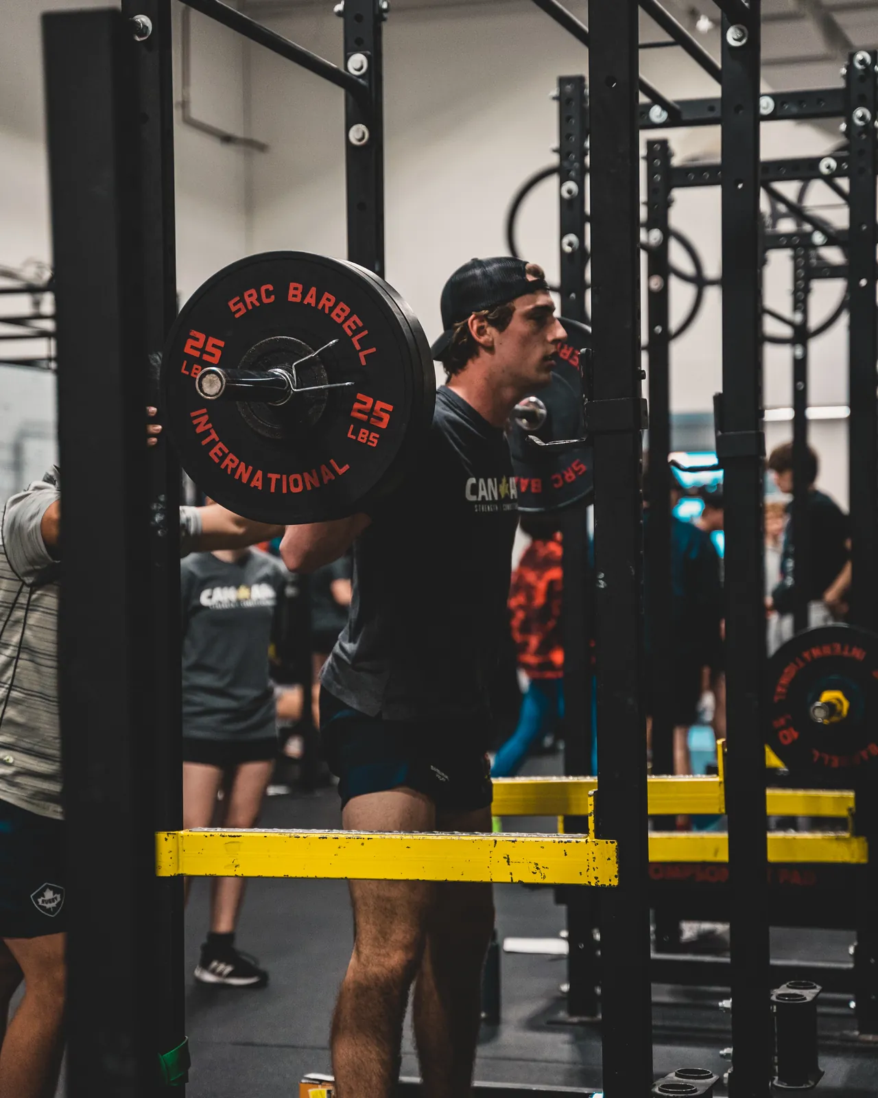 An athlete in a CANAM shirt squatting with a barbell in the rack