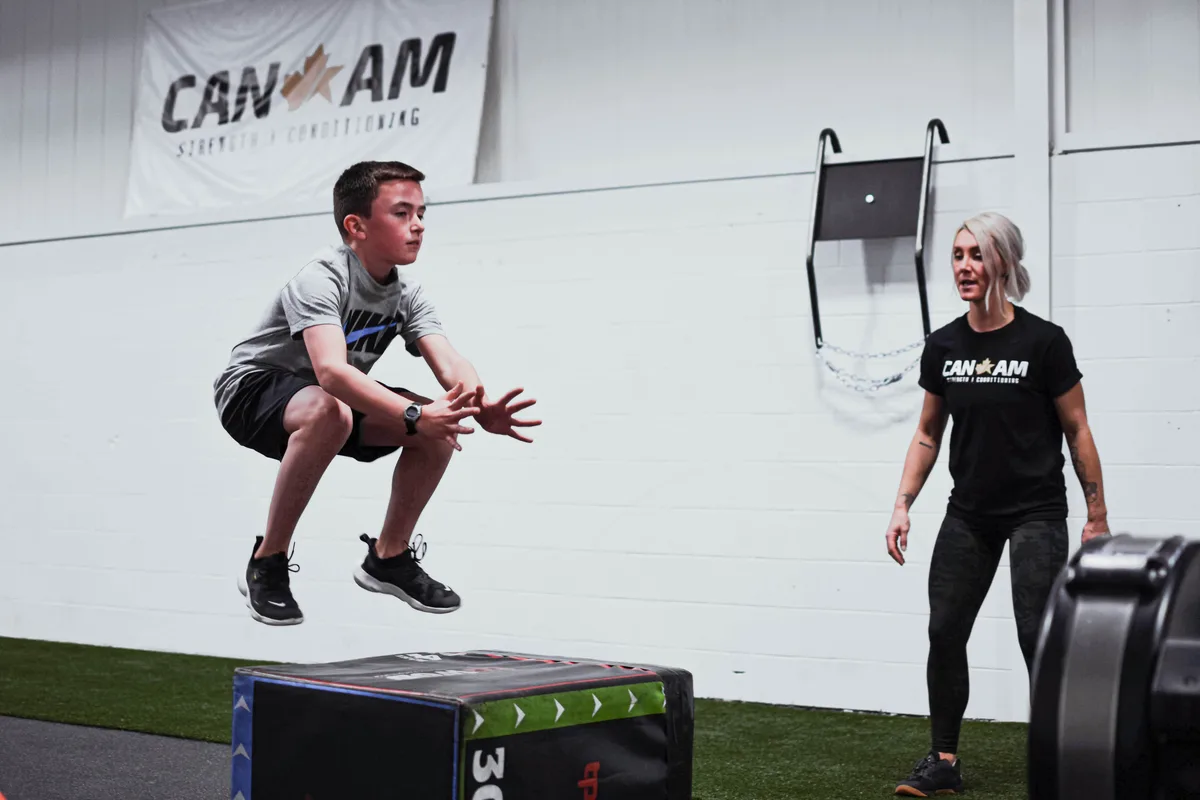 A youth athlete performing a box jump with a CANAM coach watching